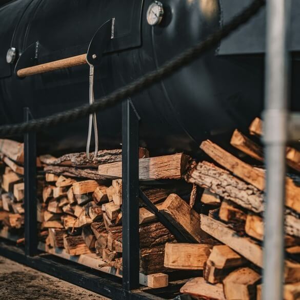 Assorted BBQ smoking woods with grill and smoked meats in the background, showcasing different wood types for BBQ and smoking.